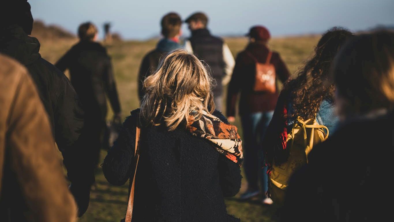 A group of people walking outdoors on a grassy area, some wearing backpacks, with a clear sky in the background.